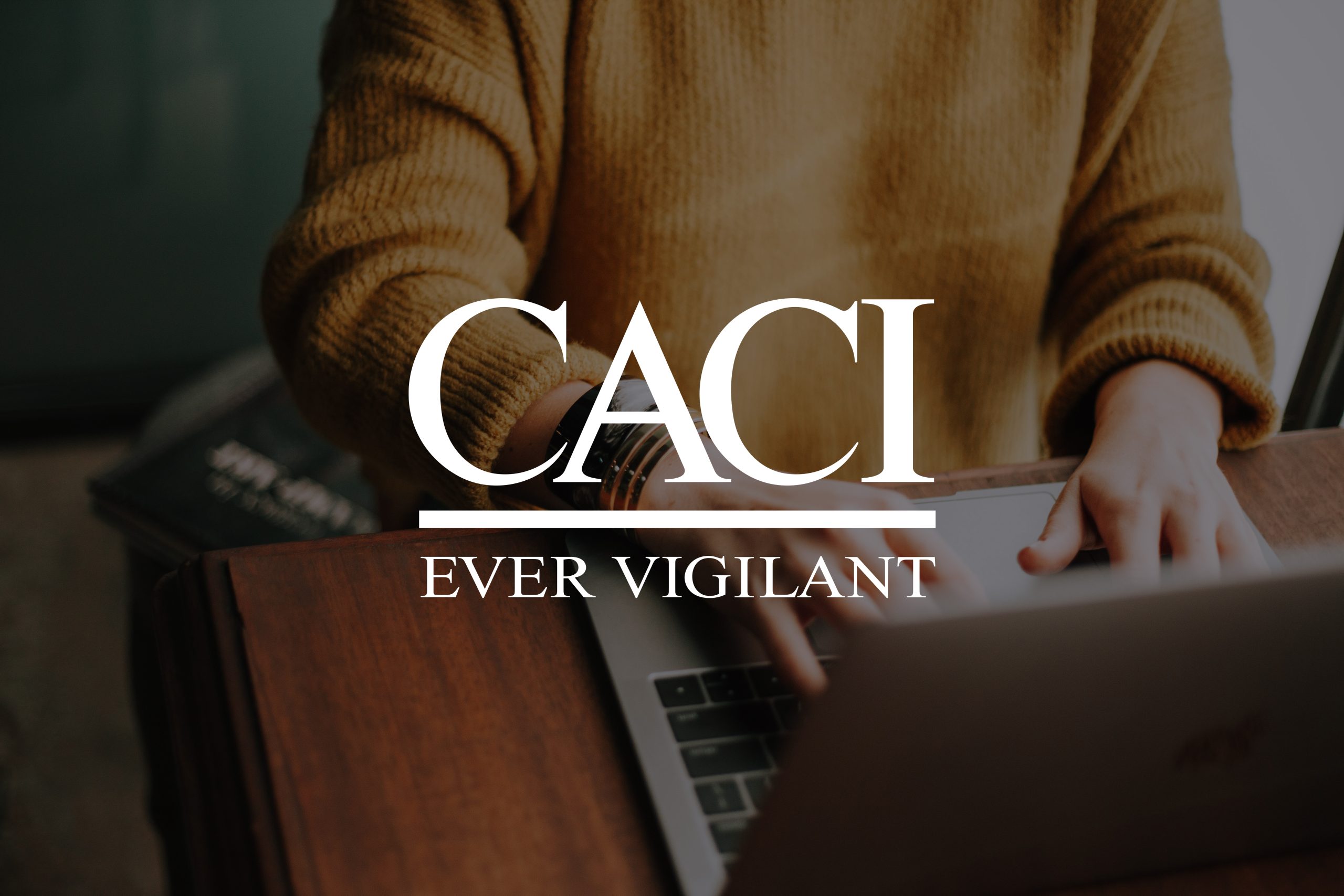 An employee wearing a yellow jumper, typing on a laptop, with the CACI logo displayed on top